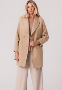 Beżowy płaszcz z wełny o podwójnym zapięciu z klapami i dwoma guzikami z przodu. Posiada boczne kieszenie oraz luźny krój. Teksturowane wykończenie.