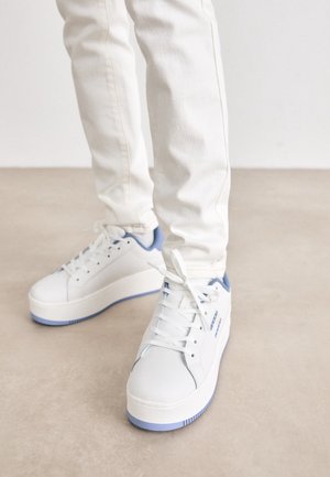 Zapatillas deportivas blancas con un acabado suave, con detalles en azul claro en el talón y la suela, y cordones planos blancos. Uso con pantalones de color claro.