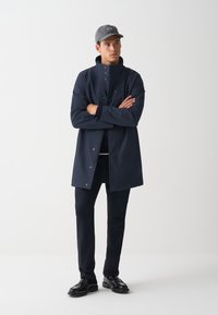 Marineblauwe waterdichte jas, staande kraag, drukknoopsluitingen, gecombineerd met donkere broek en zwarte schoenen, onder een grijze pet. Eenvoudig, functioneel ontwerp.