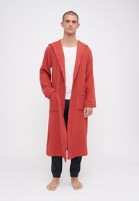 Lang, rustfarget kappe med teksturert stoff, hette og to frontlommer, båret over en hvit undertrøye og svarte joggebukser.