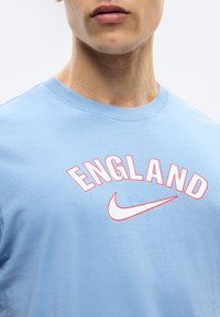 Νεαρός ενήλικας που φοράει ένα γαλάζιο μπλουζάκι με την επιγραφή "ENGLAND" και ένα λευκό λογότυπο Nike Swoosh με κόκκινο περίγραμμα στο στήθος.