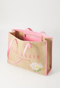 Keltainen kudottu laukku vaaleanpunaisilla nahkakahvoilla. Siinä on brodeerattu kukkakuvio ja "TED BAKER LONDON" -logo vaaleanpunaisena pinnassa.