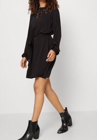 Svart klänning med spetskant i halsen, långa ärmar och bälte. Matchad med svarta ankelskängor med en texturerad yta och blockklackar.