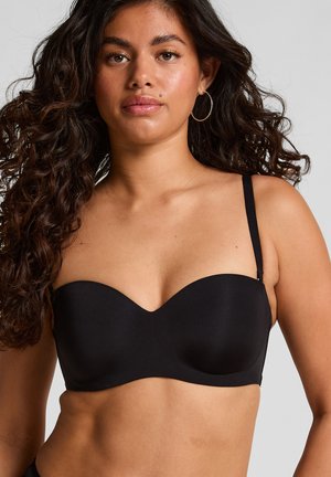 Kobieta z długimi kręconymi włosami nosząca czarny biustonosz bez ramiączek z jednym widocznym ramiączkiem na jednolitym szarym tle.