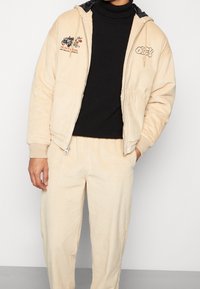 Beige kordjacka med dragkedja och svart huva, med broderade detaljer och matchande kordbyxor i avslappnad passform.