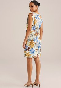 Blumenkleid mit kurzen Ärmeln in hellem Creme, verziert mit blauen und braunen Blättermustern. Tailliert, knielang, kombiniert mit hellen Schuhen.