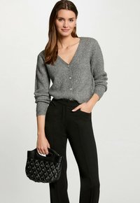 Grijze gebreide cardigan met een V-hals en knoopsluiting, gecombineerd met zwarte, op maat gemaakte broek. Model houdt een gestructureerde zwarte handtas met een handvat vast.