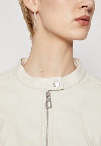 Veste en cuir blanc avec un col haut, fermeture éclair argentée et bouton-pression. Présente une texture lisse et des détails de couture.
