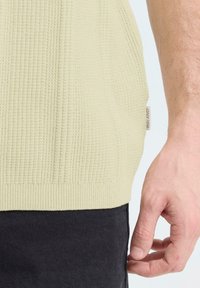 Beige gebreide truizoom met een klein "Blend" label, gedragen boven zwarte broek, naast een ontspannen hand en pols.