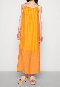 Robe maxi orange avec un décolleté froncé et des bretelles fines, présentant un tissu léger et légèrement transparent ainsi qu'une coupe ample.