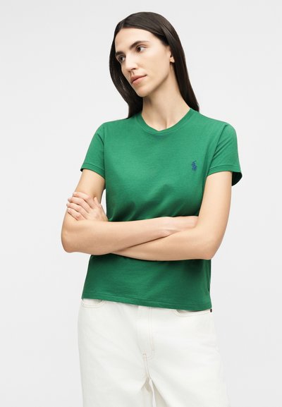 Γυναίκα με μακριά σκούρα μαλλιά που φοράει πράσινο κοντομάνικο πουκάμισο και άσπρο παντελόνι, στέκεται με τα χέρια σταυρωμένα και κοιτάζει προς το πλάι.