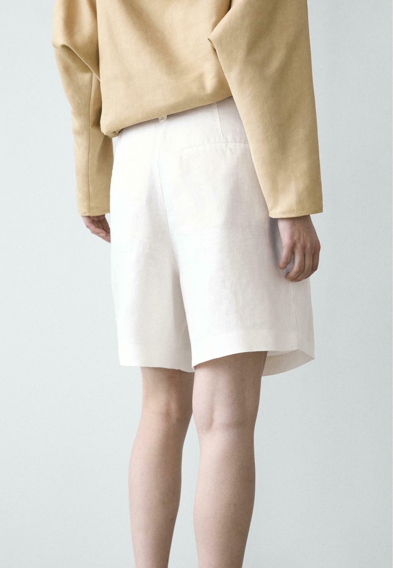 Persona che indossa una maglia beige a maniche lunghe e pantaloncini bianchi a vita alta, in piedi contro uno sfondo chiaro e uniforme.