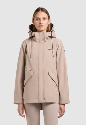 Woman wearing beige hooded waterproof jacket with front pockets and black drawstrings, standing against a plain light background.