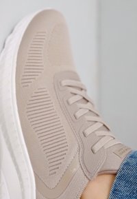 Sneaker de color beige claro con un upper de tela tejida, que presenta rayas texturizadas y una suela de caucho blanca. Diseño con cordones y un branding mínimo.