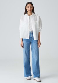 Camicia bianca leggera con bottoni e due tasche, indossata sopra una t-shirt bianca aderente. Abbinata a jeans a vita alta con gamba larga blu e sneakers bianche.