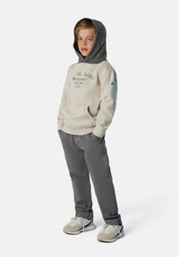 Sudadera beige con capucha gris y texto impreso, combinada con pantalones grises y zapatillas ligeras. Cuenta con un bolsillo canguro y mangas acampanadas.