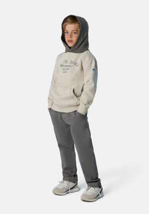 Sudadera beige con capucha gris y texto impreso, combinada con pantalones grises y zapatillas ligeras. Cuenta con un bolsillo canguro y mangas acampanadas.