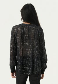 Blusa nera trasparente con motivo a pois dorati, scollatura arricciata e polsi elasticizzati. Tessuto fluido e vestibilità morbida; dettagli di design sottili.