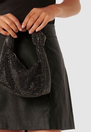 Borsa nera con una maniglia annodata, ricoperta di piccoli cristalli scintillanti, tenuta da mani davanti a una gonna di pelle nera.