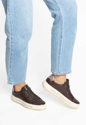 Person wearing light blue jeans and dark brown leather sneakers with white soles, standing on tiptoes against white background.