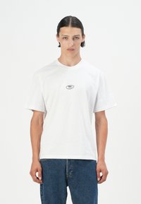 White cotton t-shirt with a crew neckline and short sleeves, featuring a small black logo on the chest. Worn with blue jeans.