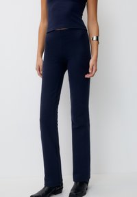 Tmavě modré pruhované dlouhé kalhoty s přiléhavým střihem, vysokým pasem a texturovanou látkou, spárované s odpovídajícím topem bez rukávů a černými botami.