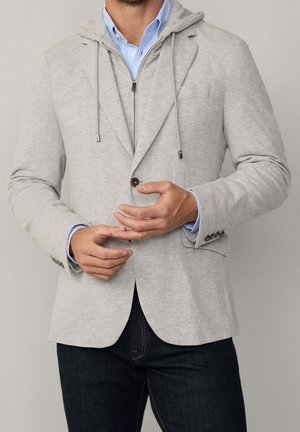 Mann trägt einen hellgrauen Blazer über einem grauen Zip-Hoodie und einem blauen Hemd mit Knöpfen sowie dunkle Jeans, passt einen Ärmelaufschlag an.