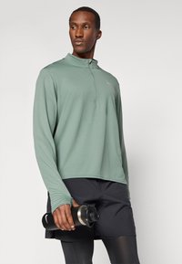 Hellgrünes langärmliges Sportoberteil mit einem Viertelreißverschluss-Design, weicher Textur und Logodetail, kombiniert mit schwarzen Shorts und einer schwarzen Wasserflasche.