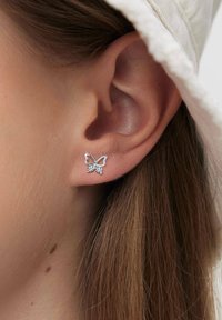 Srebrna kolczyk w kształcie motyla z przezroczystym akcentem kamienia szlachetnego, o otwartej konstrukcji. Noszony na płatku ucha, ukazuje finezyjne detale i fakturę.
