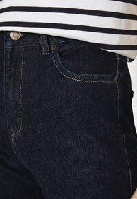 Jeans de ganga azul escuro com costura contrastante amarela, design de cinco bolsos e fecho metálico na cintura.