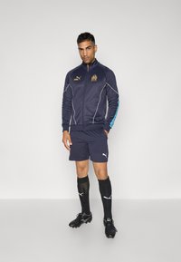 Námornícky tréningový oblek s bundou na zips a šortkami. Obsahuje značku Puma, zlaté akcenty a biely lem. Čierne futbalové kopačky s kontrastnými ponožkami.