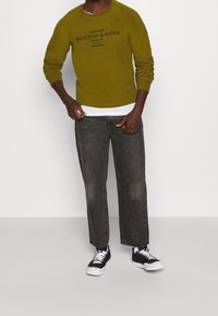 Olivengrøn sweatshirt med sort tekst, kombineret med grå denimjeans. Outfit'et fuldendes med sorte sneakers med hvide såler.