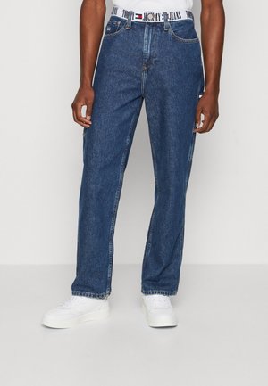 Mężczyzna w niebieskich prostych jeansach z białym paskiem Tommy Jeans i białymi trampkami na jednolitym tle.
