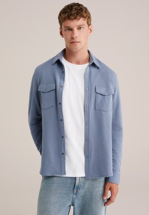 Noor mees lühikeste lokkis juustega, seljas helesinine nööbitav särk valge t-särgi ja helesiniste teksadega, seisab beežika tausta ees.