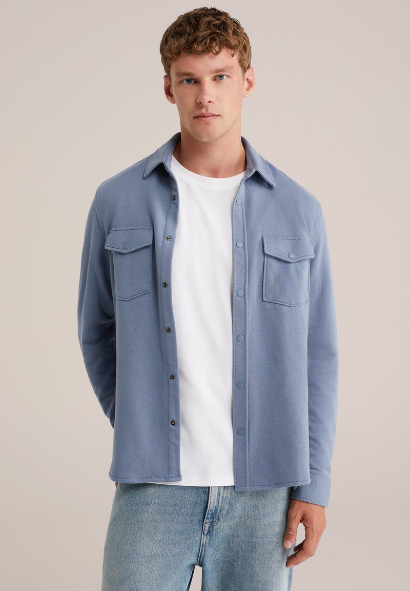 Mladý muž s krátkými kudrnatými vlasy, oblečený v světle modré košili na knoflíky přes bílé tričko a světle modré džíny, stojící na béžovém pozadí.