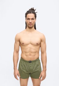 Homme musclé avec dreadlocks portant un boxer BOSS vert olive, debout devant un fond blanc uni.