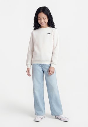 Pige med langt sort hår iført en hvid Nike-sweatshirt, lyseblå jeans med brede ben og hvide sneakers, stående og smilende, kigger ned.