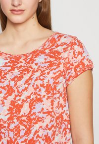 Femme portant un haut à manches courtes avec un col rond et un motif abstrait rouge, rose et blanc, montrée du menton jusqu'au bras supérieur.