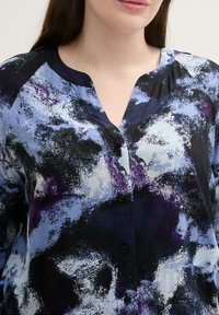 Hemdbluse mit abstrakten Mustern in Blau, Schwarz und Weiß. Weicher Stoff mit V-Ausschnitt und langen Ärmeln. Sichtbare schwarze Knöpfe.