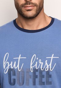 Blue cotton t-shirt with a crew neck. Features white text saying "but first COFFEE" in varying fonts and sizes. Smooth texture.