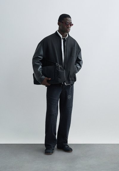 Μαύρο μπουφάν bomber με δερμάτινα μανίκια, λευκό πουκάμισο, σκισμένο σκουρόχρωμο τζιν και μαύρη τσάντα crossbody. Αξεσουάρ περιλαμβάνουν μικρά κόκκινα γυαλιά ηλίου.