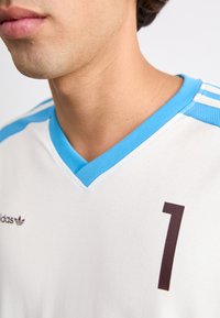 Maillot de sport blanc avec un col en V bleu clair. Présente le numéro "1" en marron et trois rayures bleu clair sur chaque épaule.