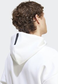 Sweat à capuche blanc en tissu texturé avec une étiquette noire contrastante au dos de la capuche. Design simple avec une coupe décontractée.