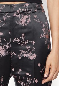 Sorte satinbukser med et delikat pink blomster mønster, struktureret talje og sidelommer. Teksturen fremstår glat og elegant.