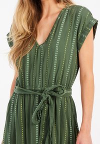 Grünes knielanges Kleid aus leichtem Stoff mit V-Ausschnitt, gefalteten Ärmeln und einem Bindegürtel. Vertikale Streifen mit weißen und gelben Punkten.