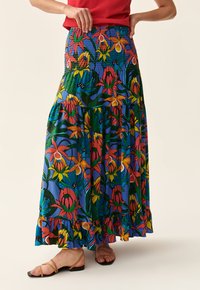 Jupe maxi florale avec taille smockée, présentant des motifs vibrants rouges, jaunes et verts sur un fond bleu ; détail de l'ourlet froncé.