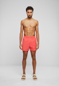Svømmeshorts i lys korallfarge, lett materiale, elastisk liv, sidelommer, med en jevn tekstur. Skoene har åpen tå-design.