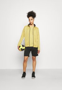 Kvinne i gul jakke og svarte shorts som holder en gul og svart håndball, stående foran en hvit bakgrunn.