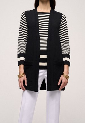 Kvinde iført sort ærmeløs cardigan med åben front over en sort og hvid stribet langærmet skjorte, hvide bukser og gyldne armbånd på begge håndled.