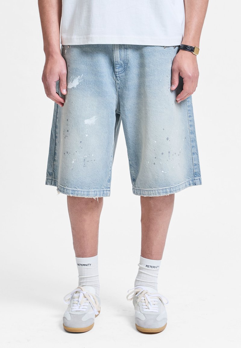 Personne portant un short en jean bleu clair usé au niveau des genoux, des chaussettes blanches avec du texte, des baskets blanches et une chemise blanche.